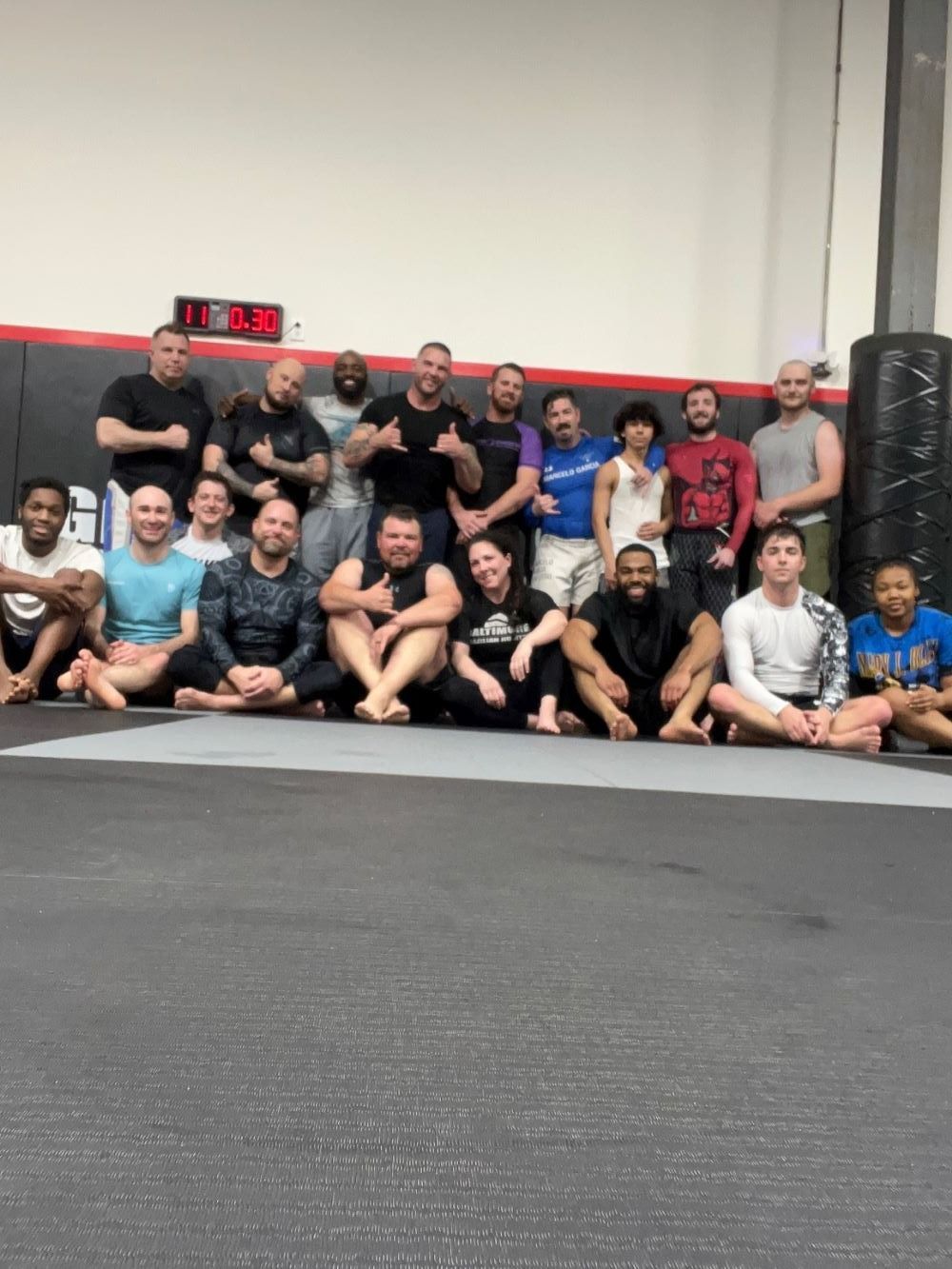 Group of people posing in a gym. Many smiling, various skin tones, seated on a mat. Digital clock visible.
