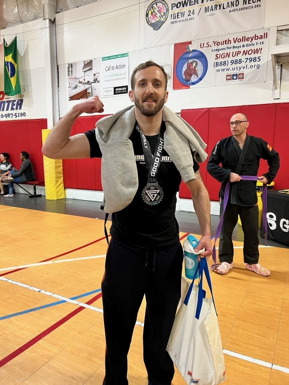 Man flexing arm, holding medal, wearing black attire, with other people in a martial arts gym.