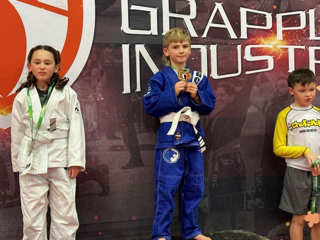 Kids on podium at grappling competition; boy with gold medal, blue gi; girl, white gi; boy, yellow shirt, gray shorts.