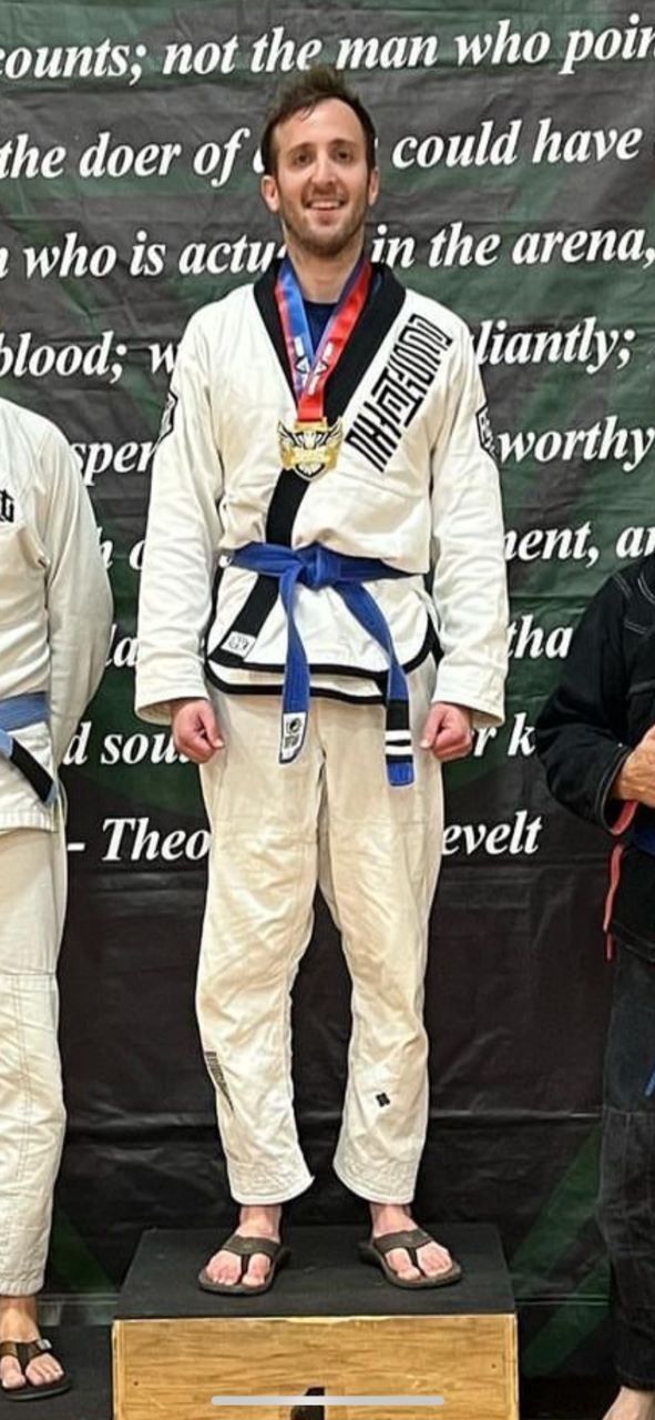 Man in a white gi with blue belt, wearing a medal, standing on a podium.