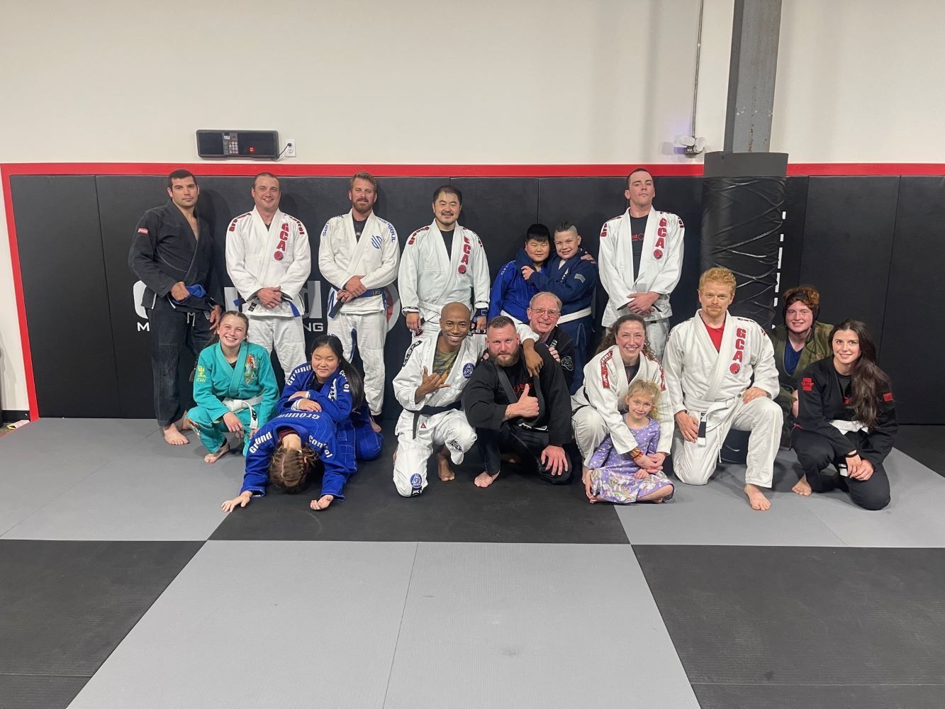 Group of people in martial arts attire posing on a mat, indoors. Smiles, various belt colors, and a celebratory feel.