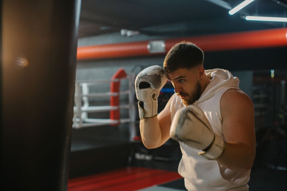 Boxer in a gym, wearing white gloves and hoodie, in a fighting stance by the ring ropes.