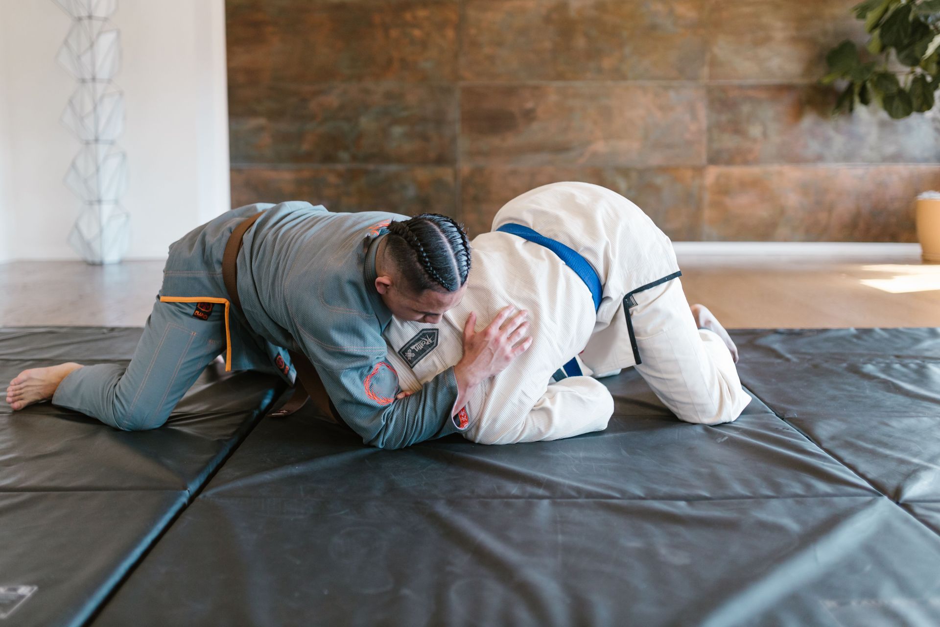 Two martial artists grappling on mats in a dojo, with two others blurred in the background.