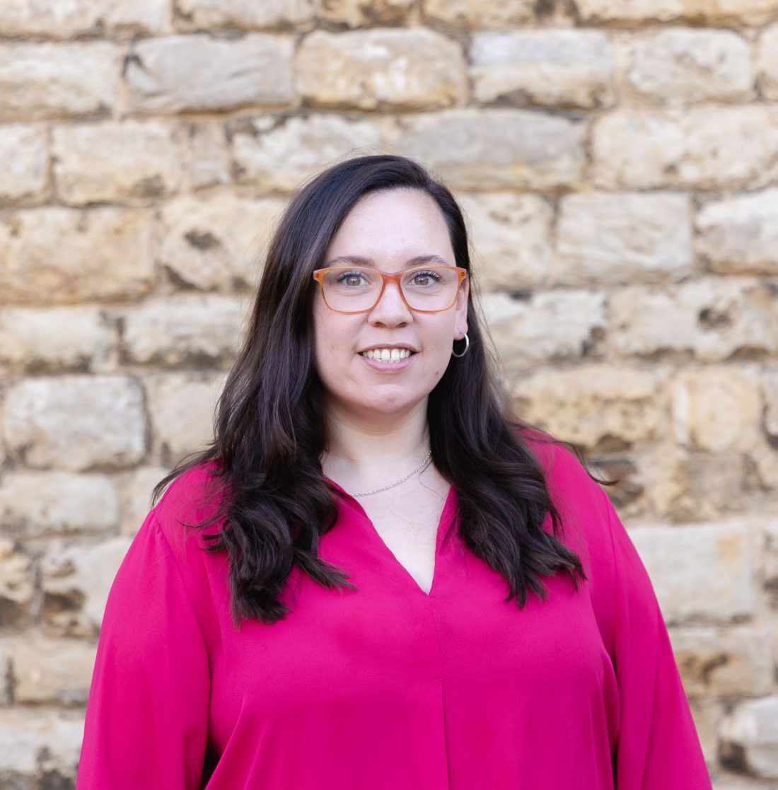 A person with long dark hair and glasses wears a pink blouse, smiling in front of a light-colored stone wall.