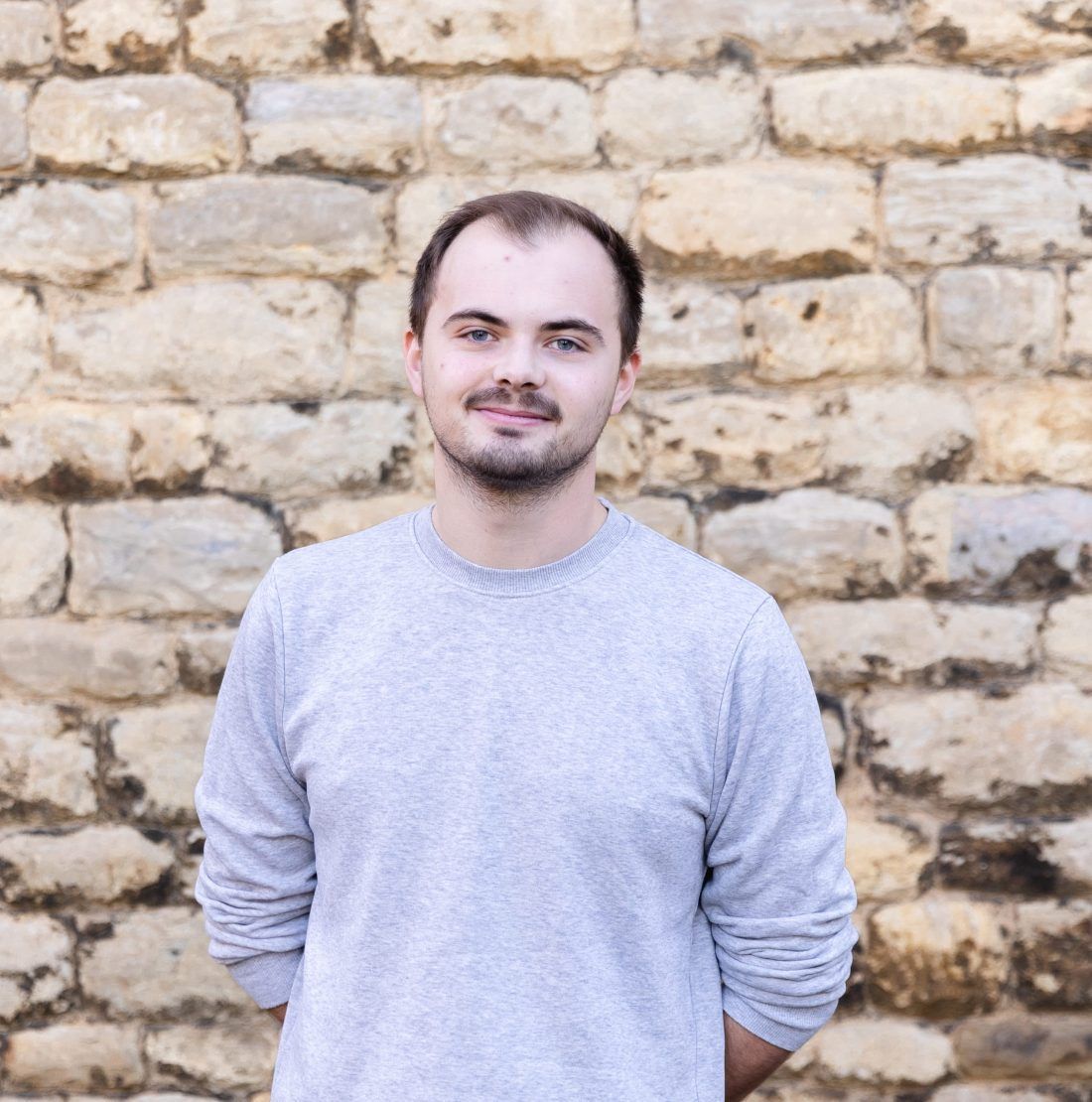 A person in a light grey long-sleeved shirt stands centered in front of a rustic, textured light-colored stone wall.