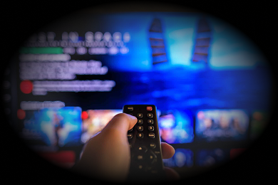 A hand holds a TV remote in front of a television screen displaying a streaming service menu with blue lighting.