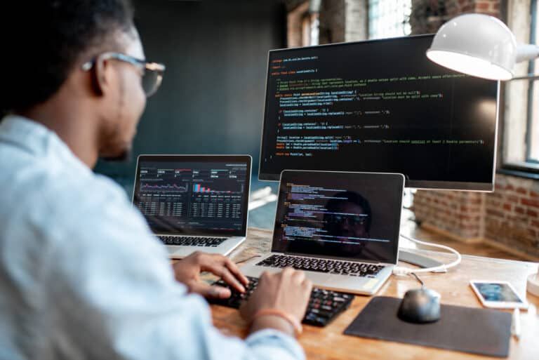 A developer working at a desk with two laptops and a large monitor displaying lines of code in a rustic brick office.