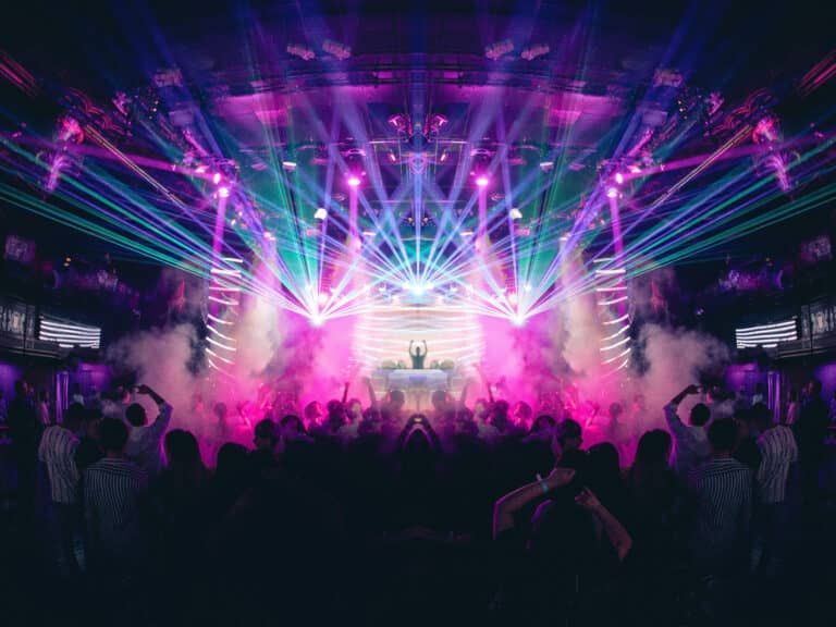 A DJ performs on stage at a crowded nightclub with vibrant laser lights, colorful stage lighting, and smoke effects.