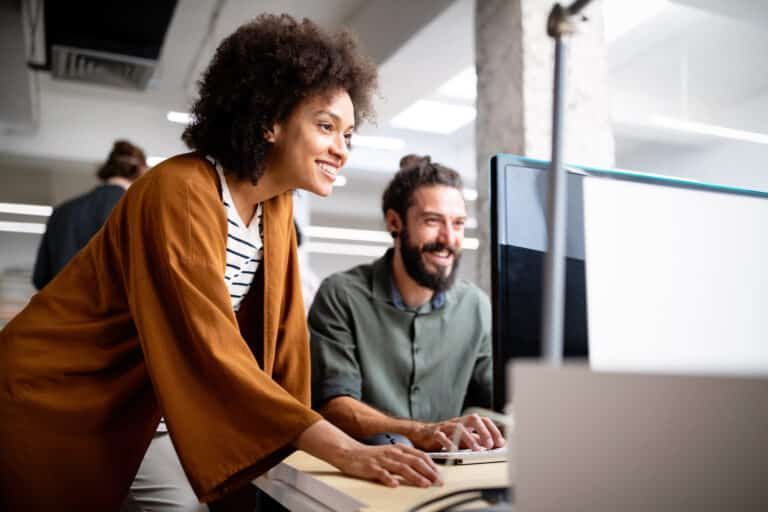 Two professionals collaborate at a desk in an office, smiling as they look at a computer monitor.
