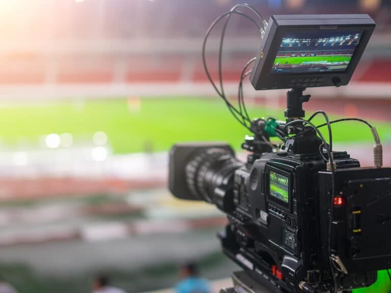 A professional broadcast video camera focused on a soccer field in a stadium, with a monitor displaying the live feed.
