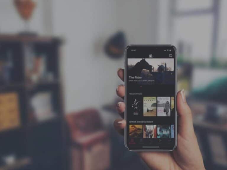 A hand holds a smartphone displaying the Apple TV app interface against a blurred indoor background.