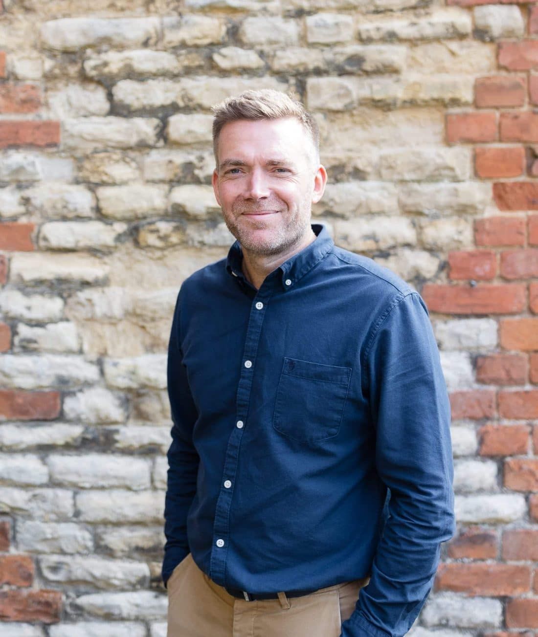 A smiling person in a blue button-down shirt and tan pants standing in front of a rustic stone and brick wall.
