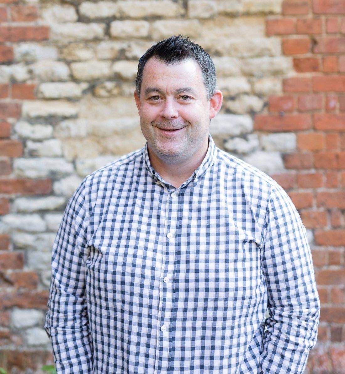 A person with short dark hair wearing a blue and white checkered shirt, standing in front of a brick and stone wall.