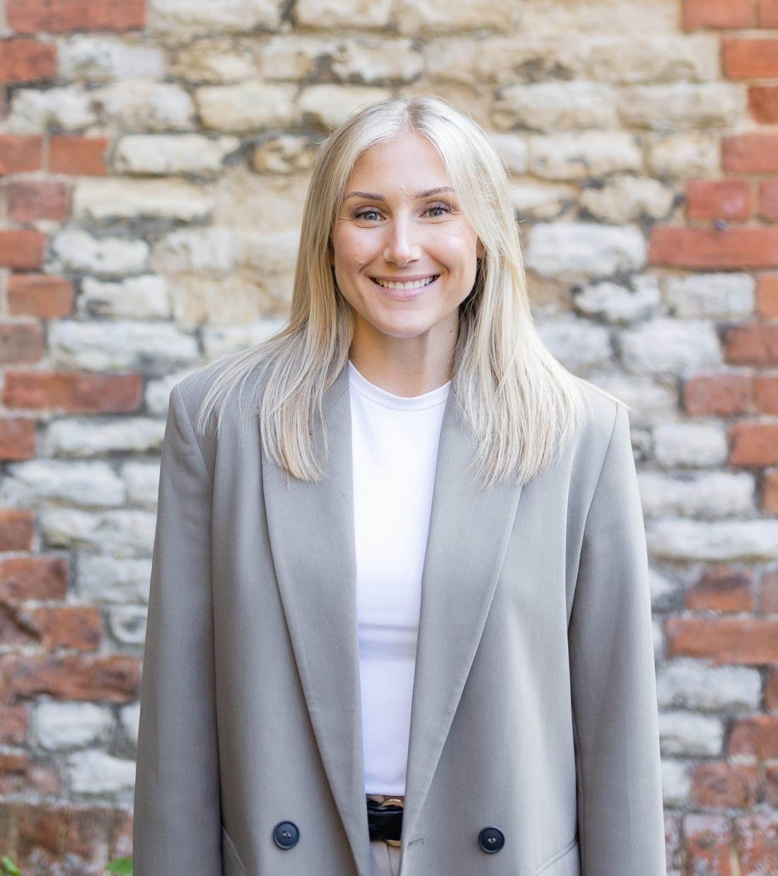A person with shoulder-length blonde hair smiles, wearing a white shirt and a grey blazer against a textured brick wall.