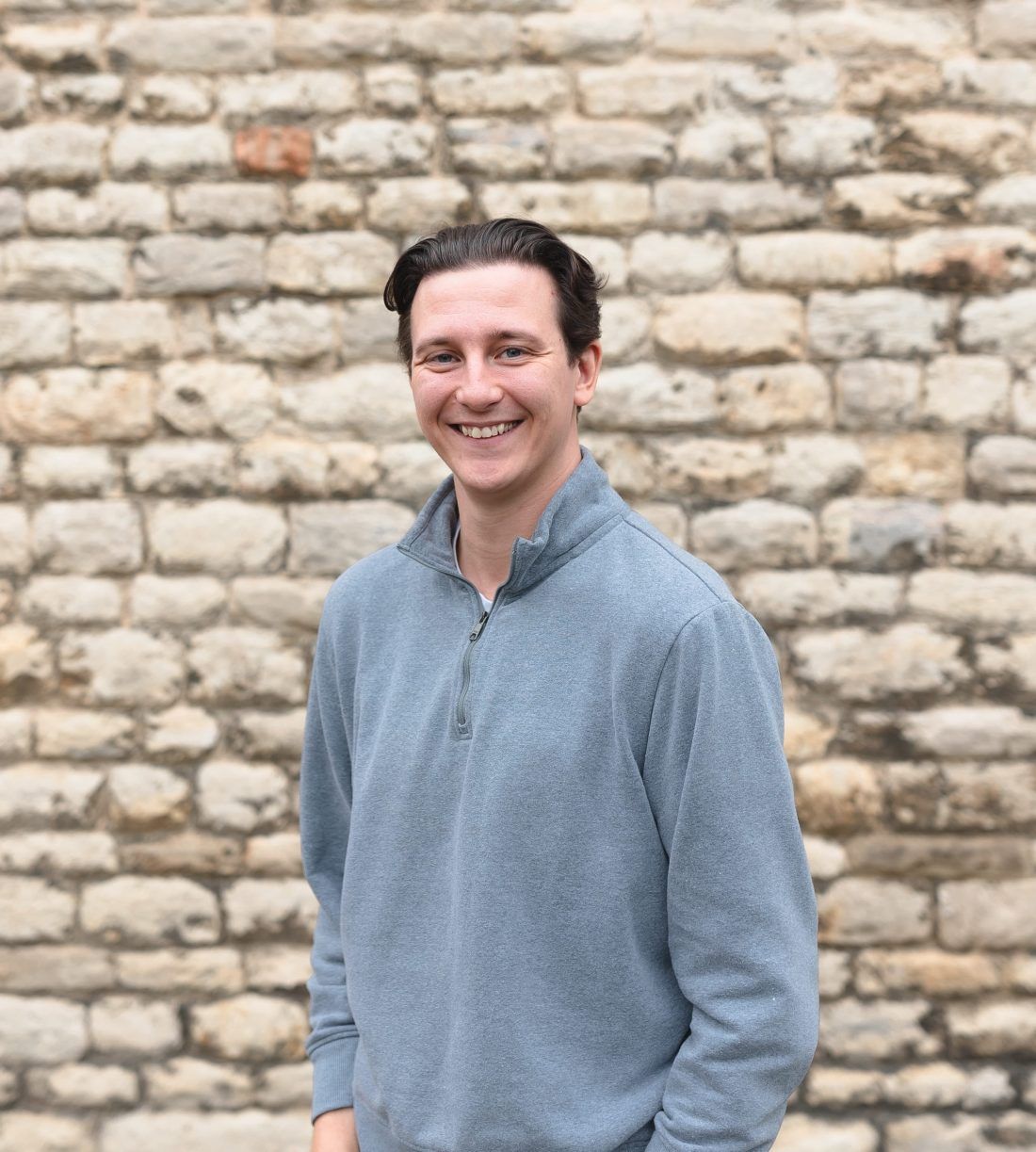 Smiling person in a blue quarter-zip sweater standing in front of a light-colored stone wall.