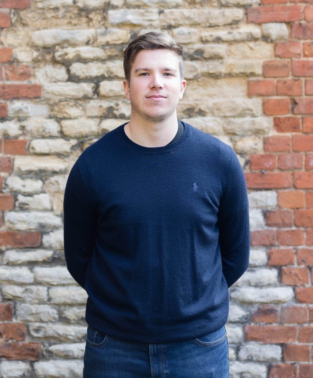 A person wearing a dark blue sweater stands against a textured wall of brick and light-colored stone.