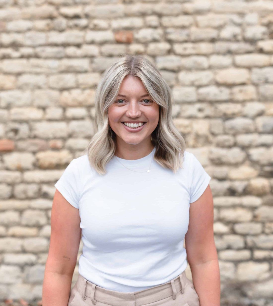 Smiling person with shoulder-length blonde hair, wearing a white t-shirt in front of a rustic stone wall.