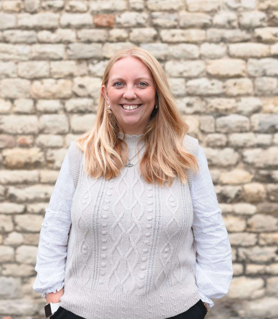 A smiling person wearing a beige cable-knit sweater over a white blouse, standing in front of a light-colored stone wall.