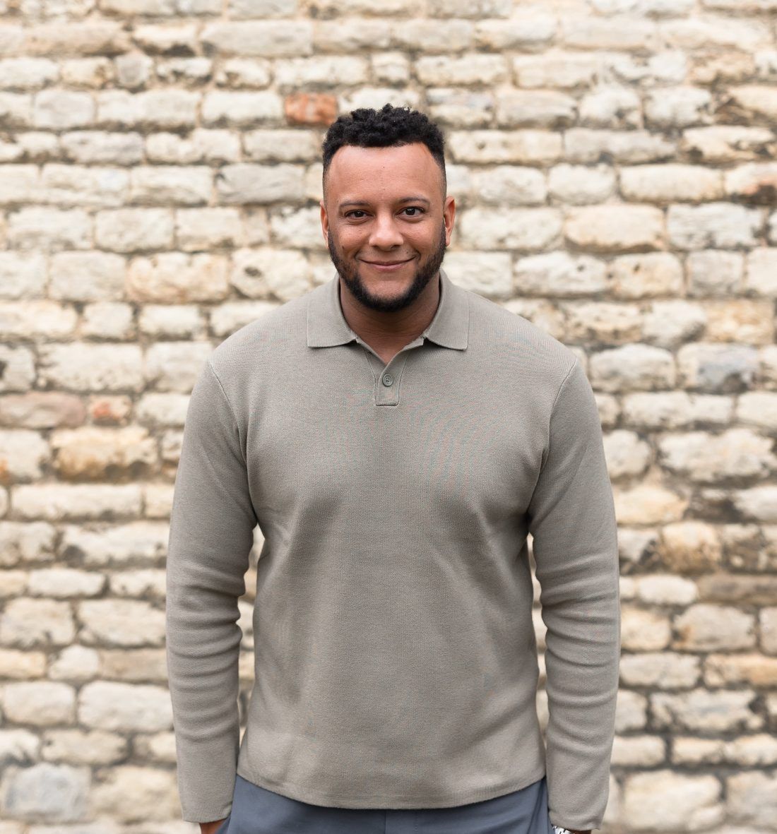 A person with a beard and short dark hair wearing a long-sleeved beige collared shirt against a stone wall background.