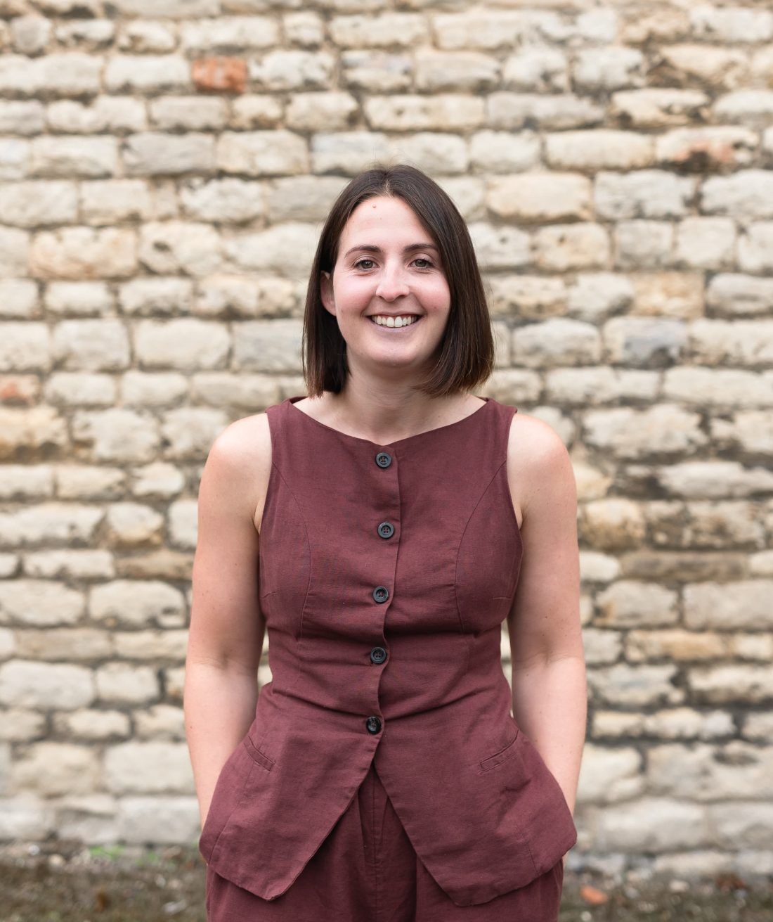A person smiles while posing in a maroon sleeveless top against a textured stone wall.