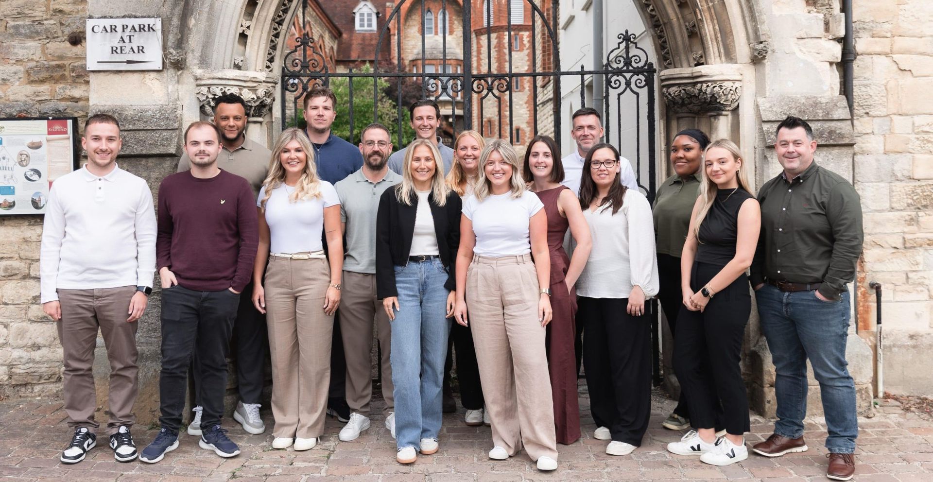 A group of fifteen people posing outdoors in front of a stone archway and metal gates, smiling at the camera.
