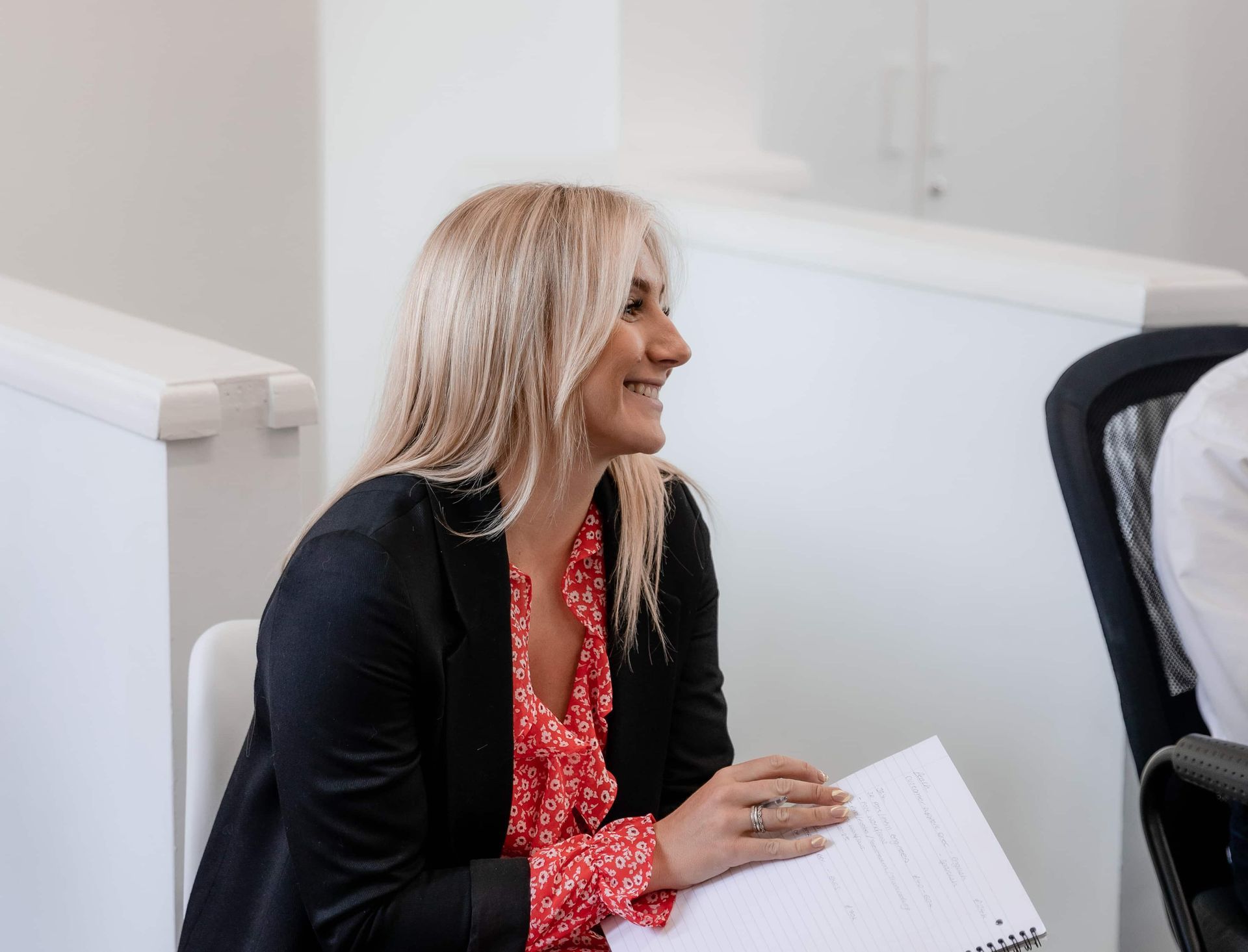 A person with blonde hair wearing a black blazer and red patterned top smiles while holding a notebook in an office.