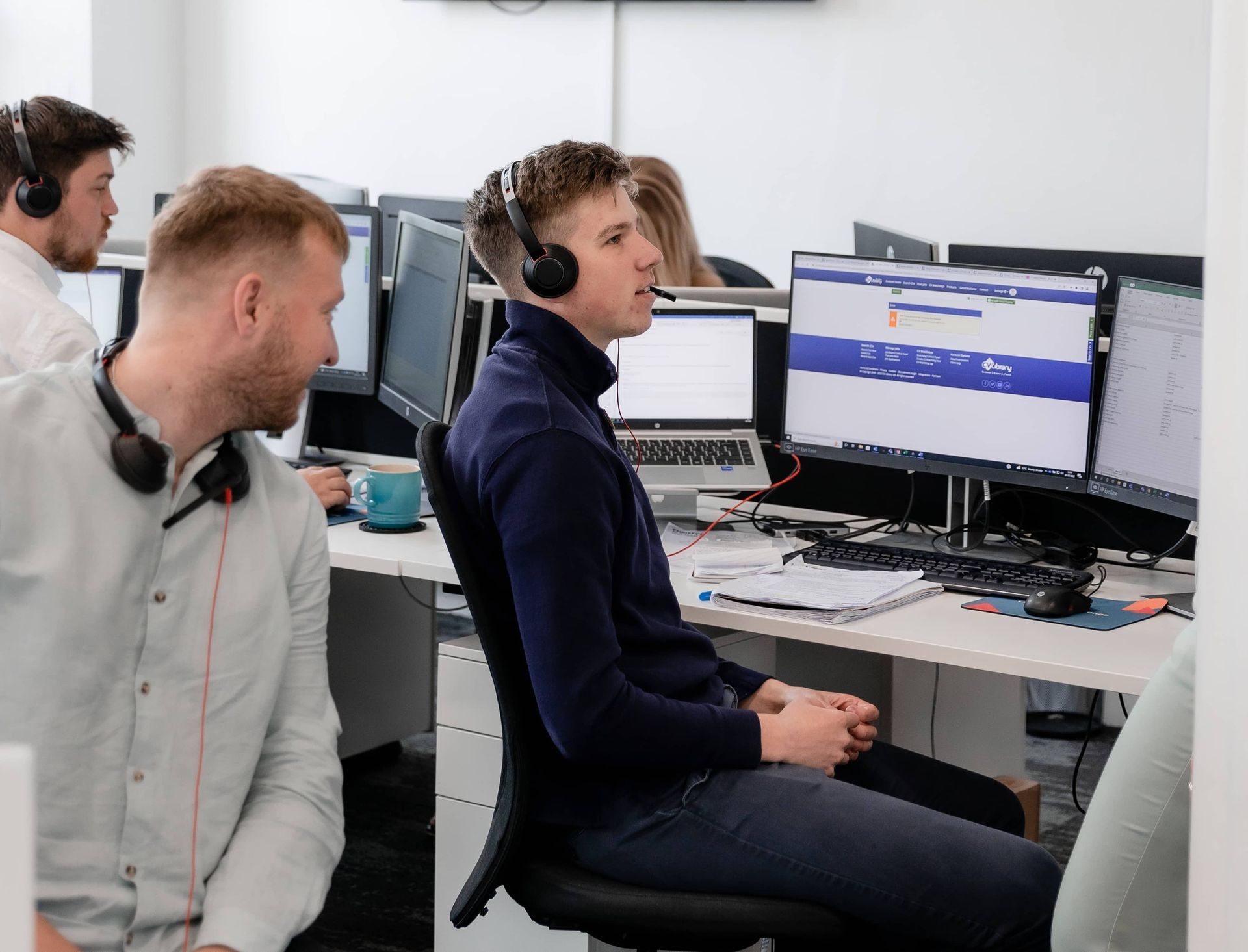 Office workers wearing headsets in an open-plan workspace, collaborating at desks with multiple computer monitors.