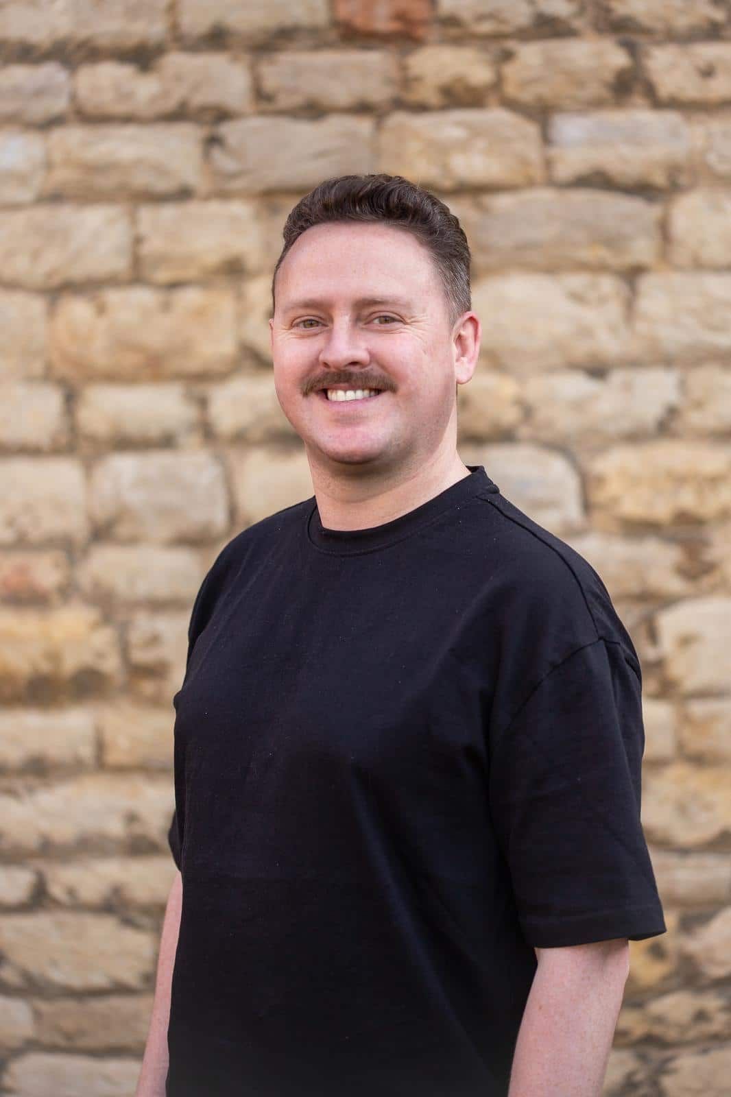 A person with a mustache smiling while wearing a black crew-neck shirt against a textured, light-colored stone wall.