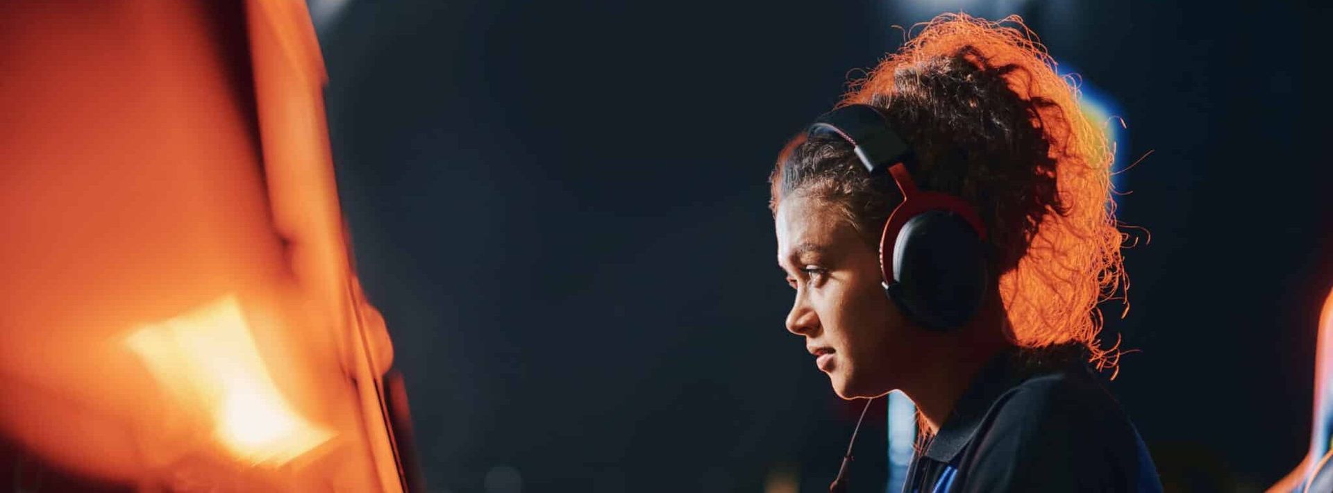 A person with curly hair wearing black headphones, intently looking at a screen in a dimly lit space with orange lighting.