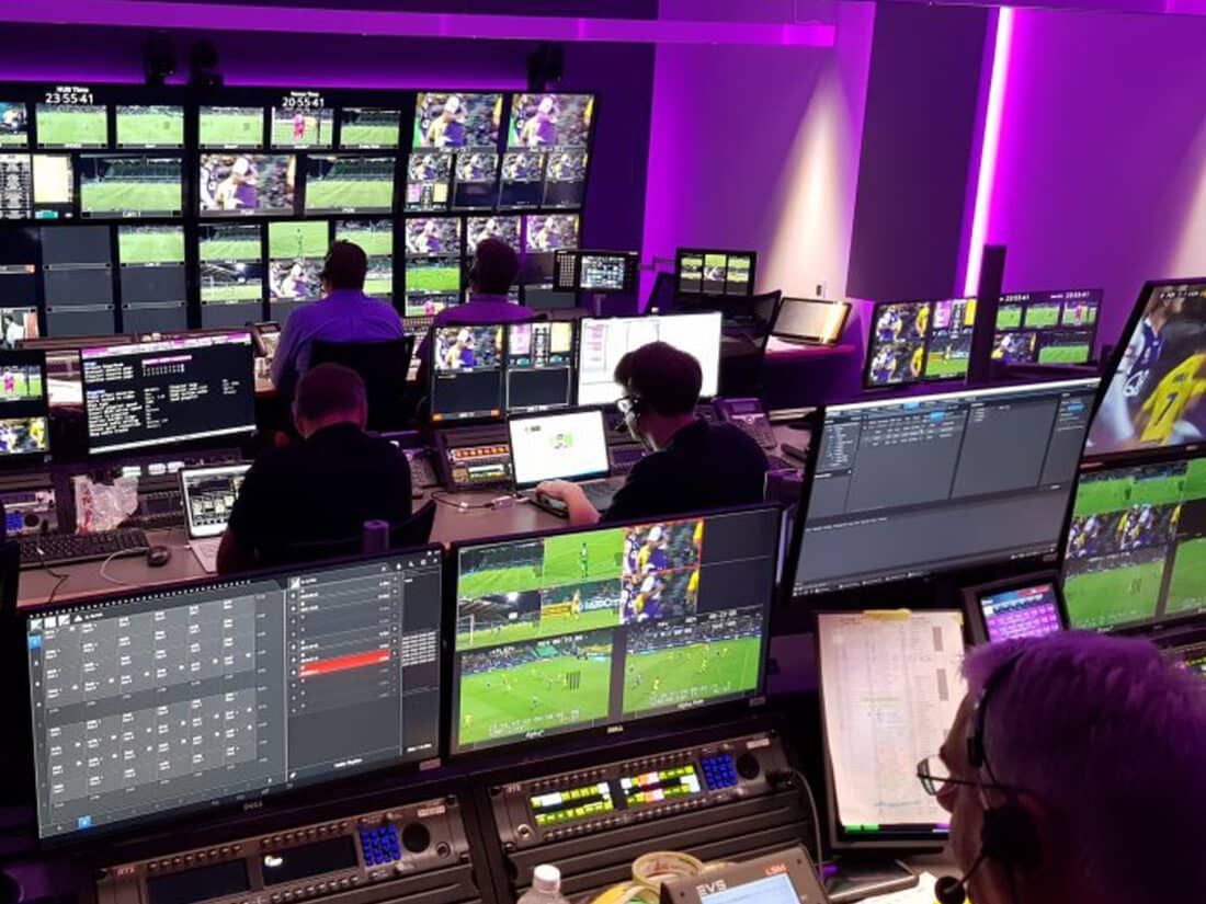Broadcast operators work in a dimly lit control room filled with multiple monitors displaying sports footage.