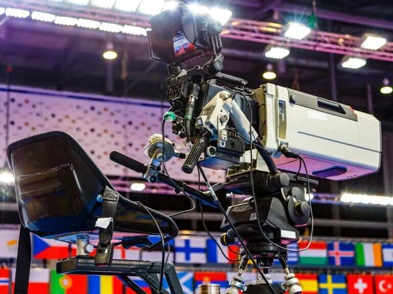 A professional television camera on a tripod, angled toward a background featuring a row of international flags.