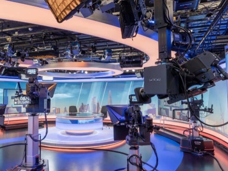 A modern, brightly lit news studio featuring a central circular desk, curved digital displays, and multiple video cameras.