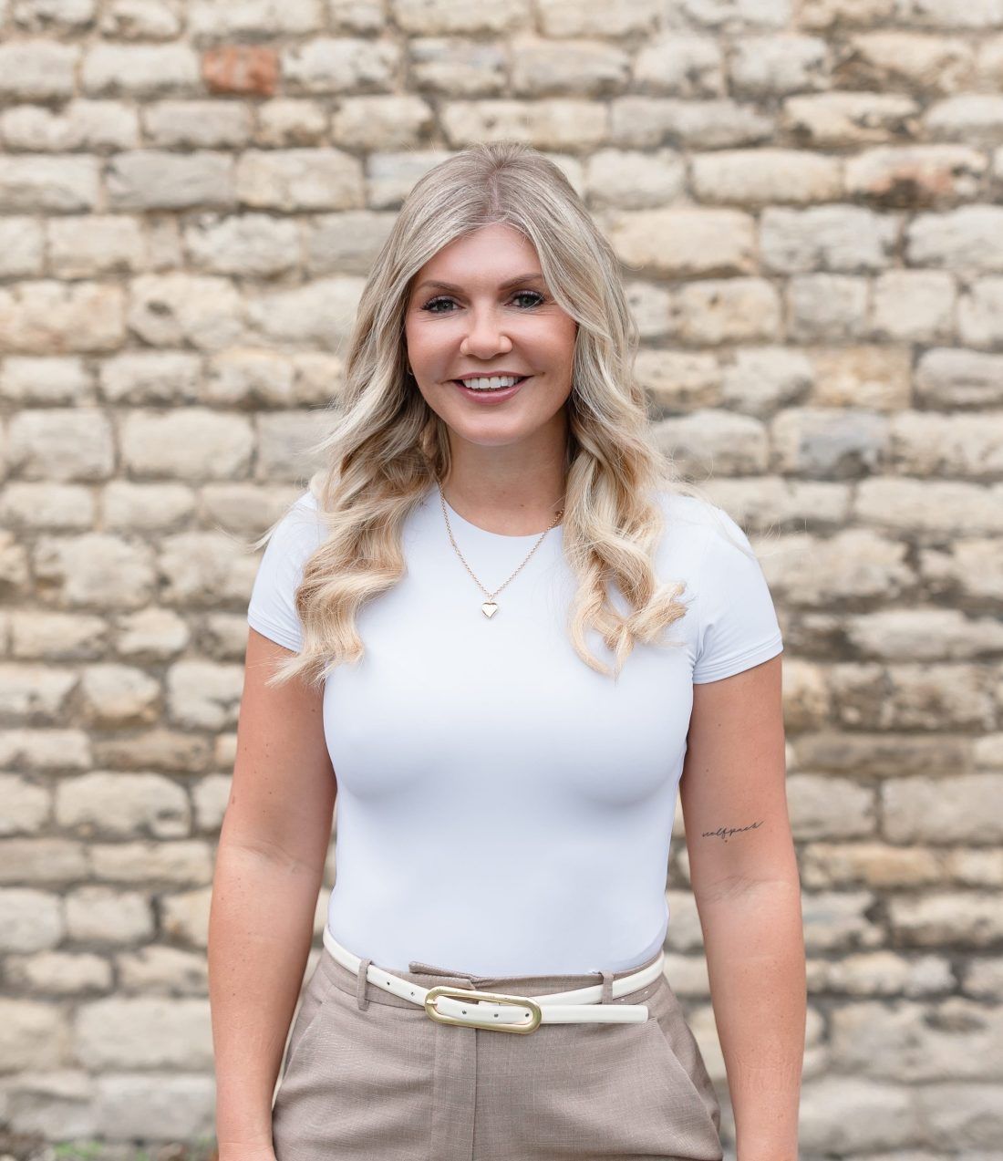 Smiling person with blonde hair wearing a light blue t-shirt and tan trousers, standing in front of a stone wall.