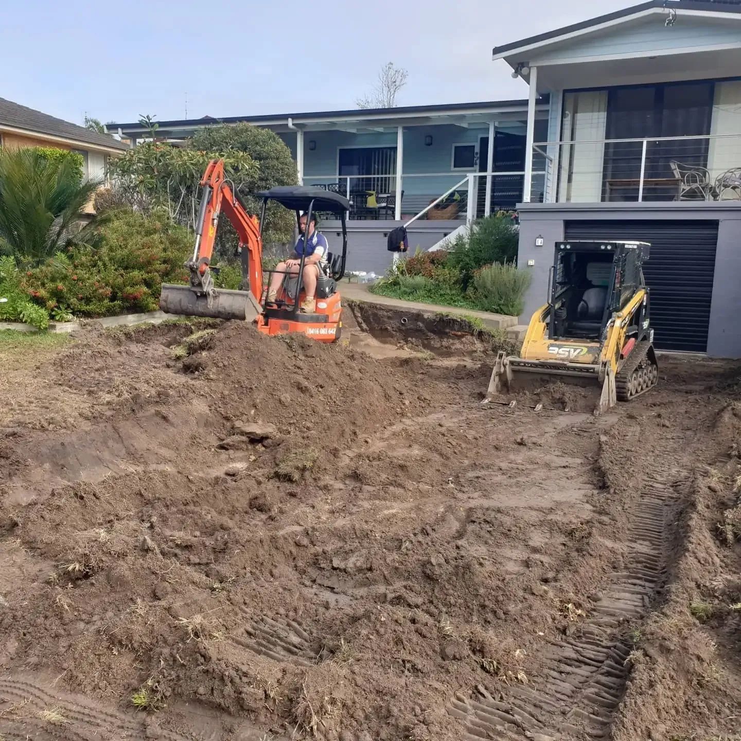 A Man Is Driving an Excavator in A Dirt Field in Front of A House — Turn It Crete in Sussex Inlet, NSW