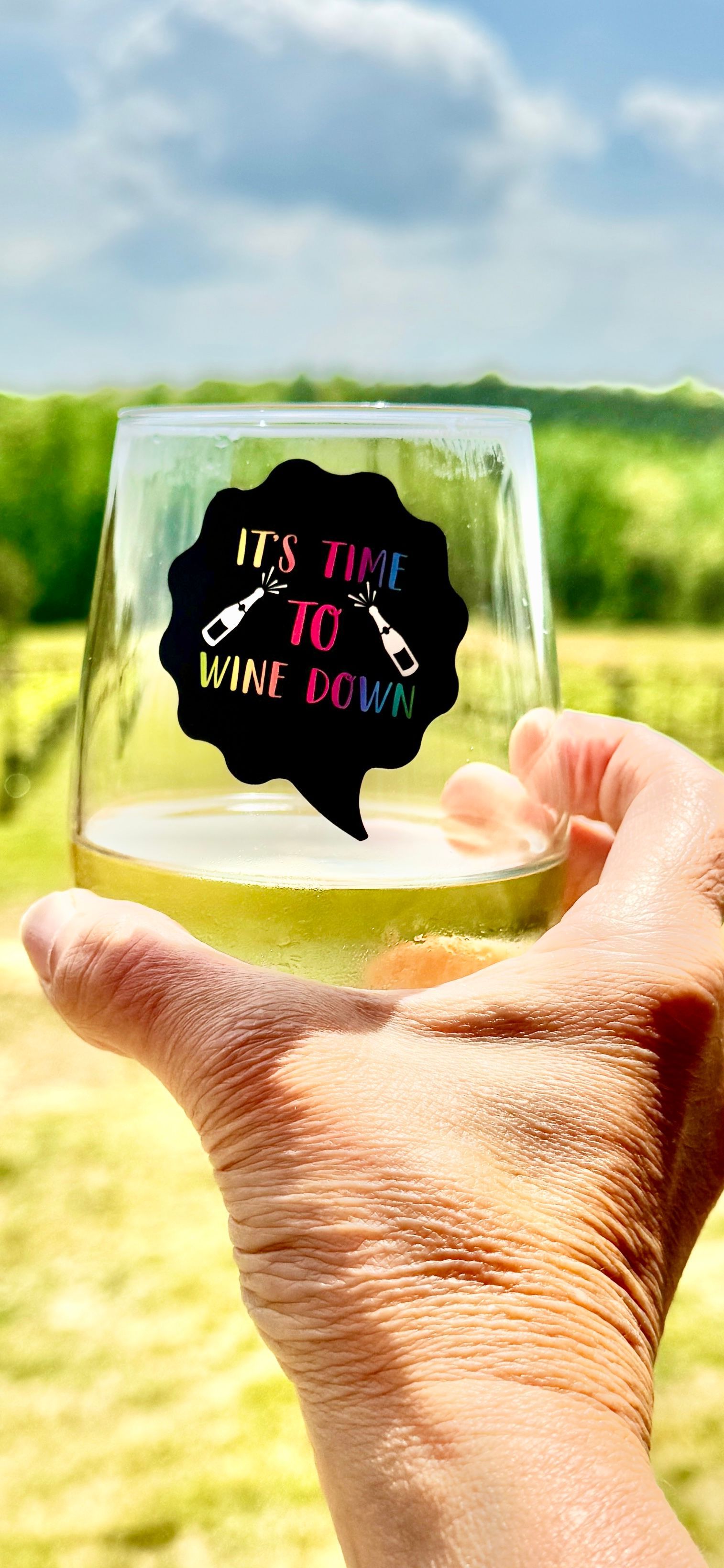 Hand holding a clear square ornament with colorful text against a blurred outdoor sky and field background