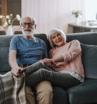 Elderly couple smiling on a couch, holding hands. Man in blue shirt, woman in pink.