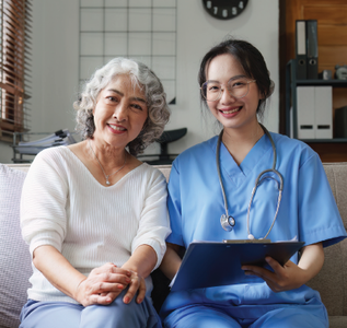Nurse with clipboard smiles next to senior patient on a sofa.
