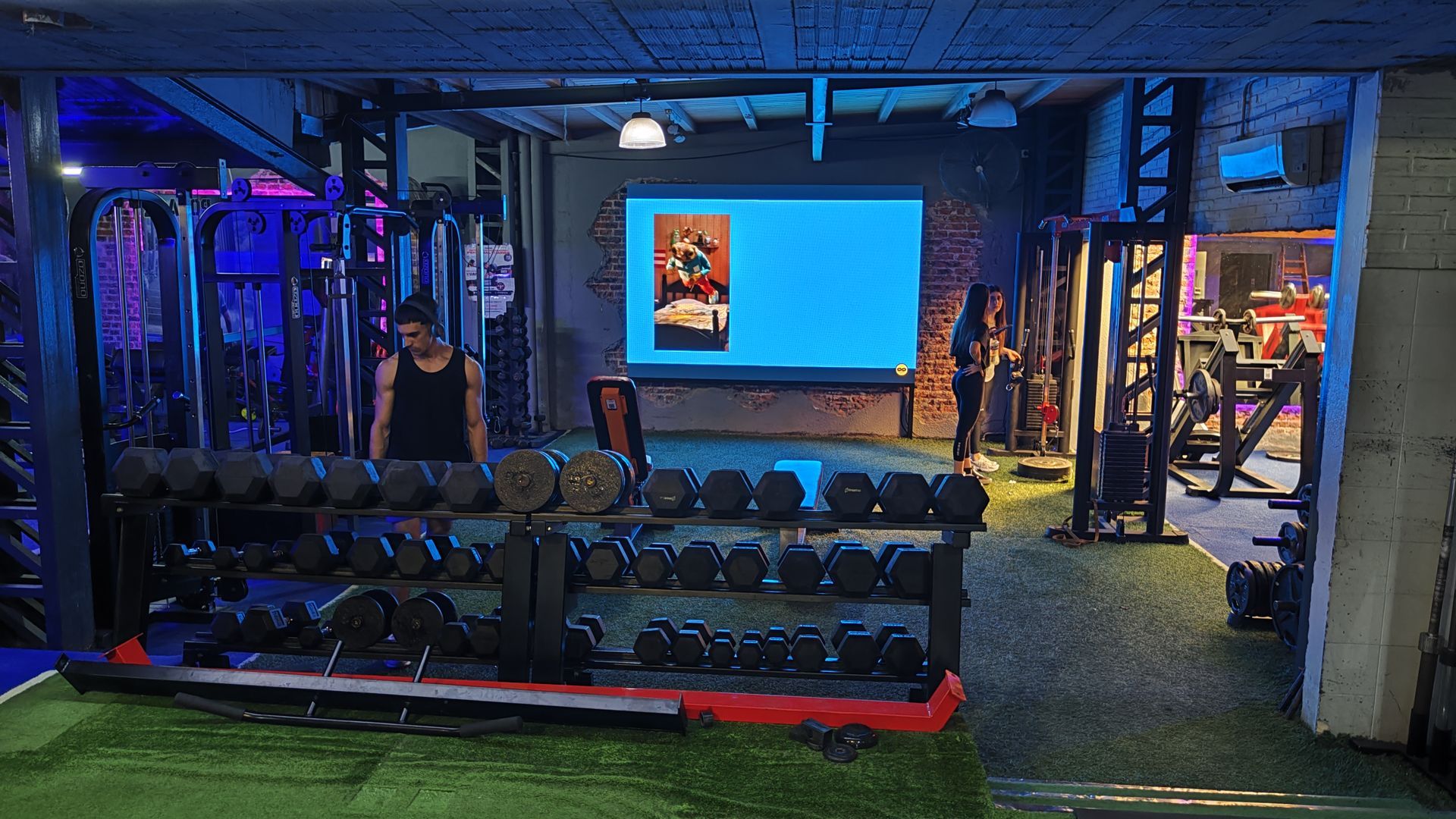 Interior de gimnasio con mancuernas, máquinas de pesas y dos personas cerca de una pantalla que muestra una imagen.