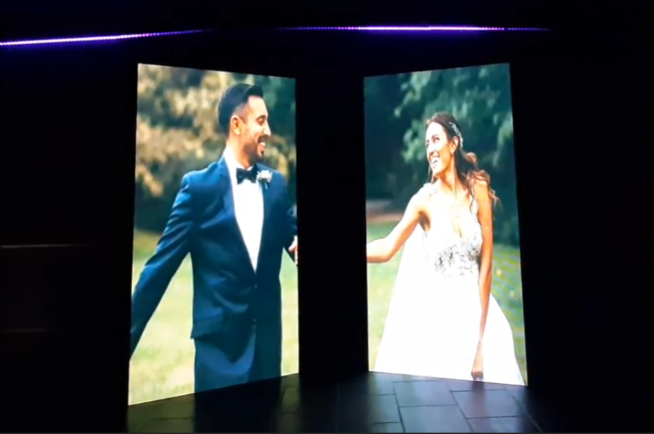 Pareja de recién casados sonriendo en pantallas, vestidos de gala, al aire libre.