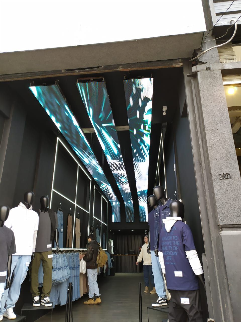 Interior de una tienda de ropa con maniquíes que exhiben jeans y camisas; pantallas digitales en el techo.