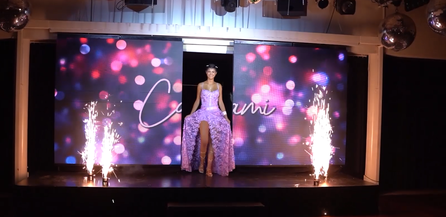 Una mujer con un vestido morado sube al escenario. Bengalas y un fondo con luces rosas y azules.