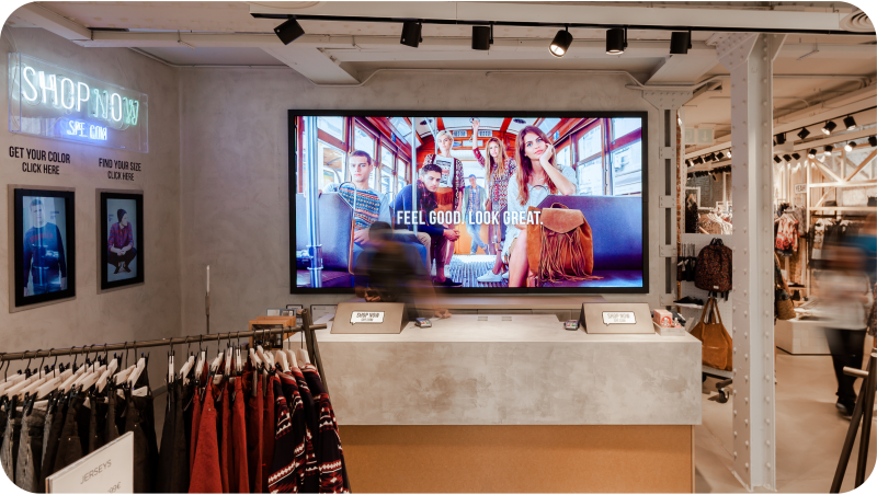 Interior de una tienda de ropa con una gran pantalla que muestra publicidad, percheros y clientes.