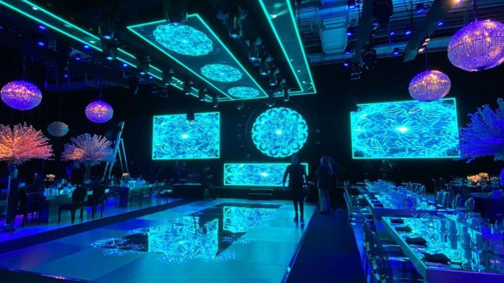 Una gala con iluminación azul y púrpura, un escenario con pantallas que proyectan arte abstracto
