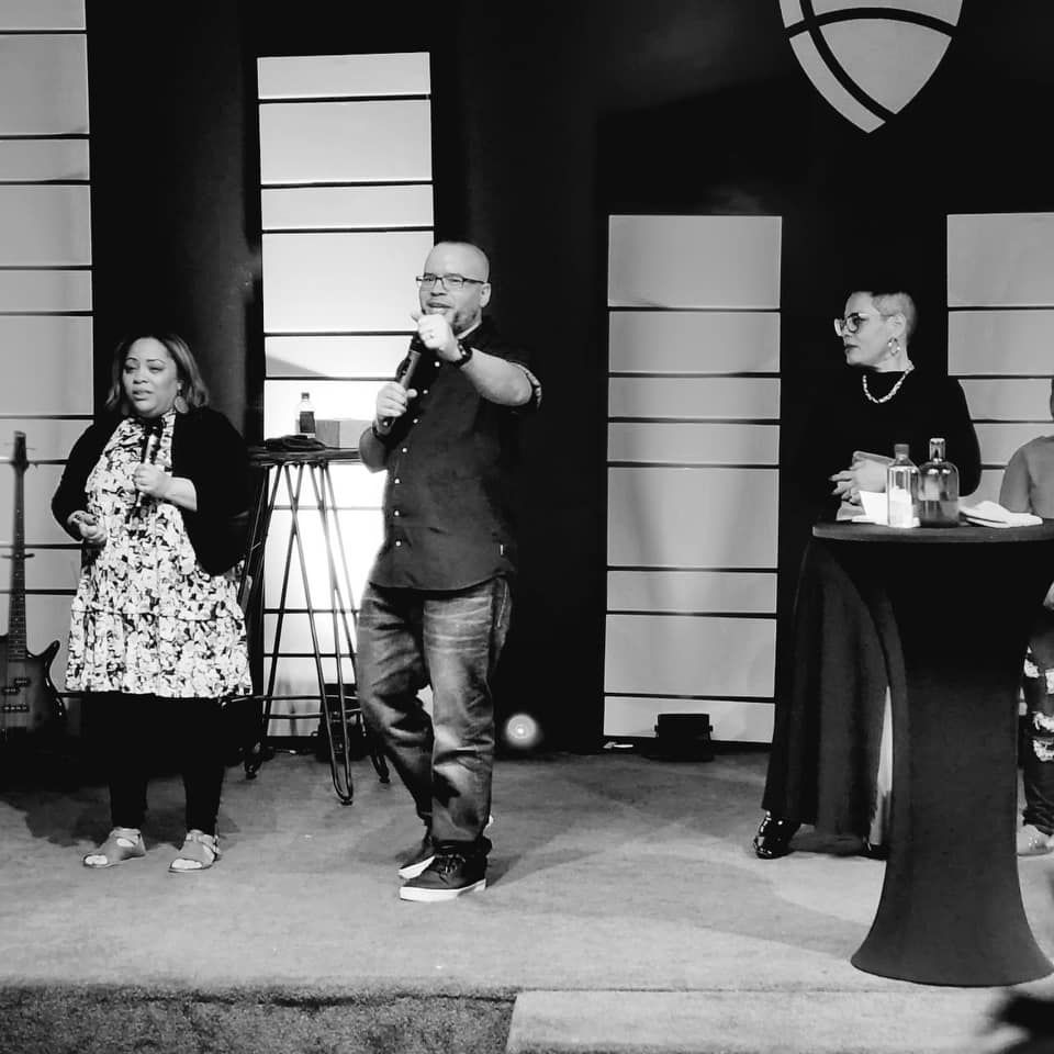 A man with a microphone speaks on stage with two women. Indoor stage. Black and white.