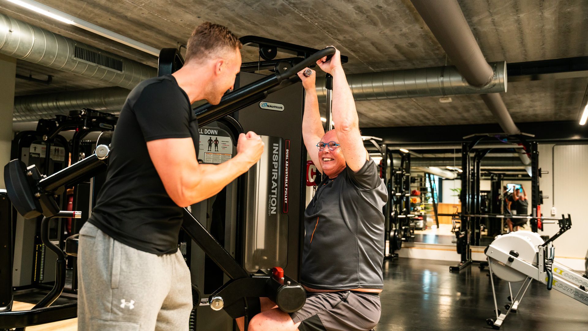 Een man wordt bijgestaan ​​door een trainer bij het gebruik van een gewichtsapparaat in een sportschool. Beiden glimlachen.