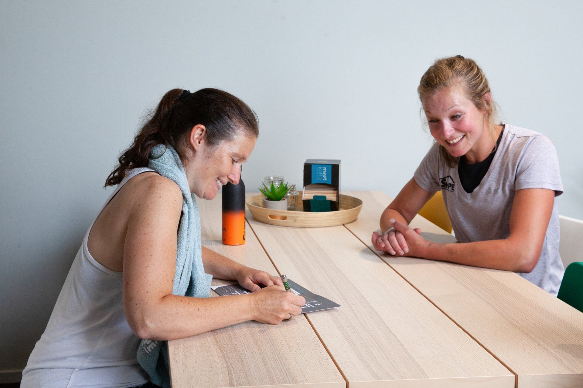 Twee vrouwen zitten aan een tafel. De ene tekent, de andere glimlacht. Op de tafel staan ​​een fles water en een dienblad.