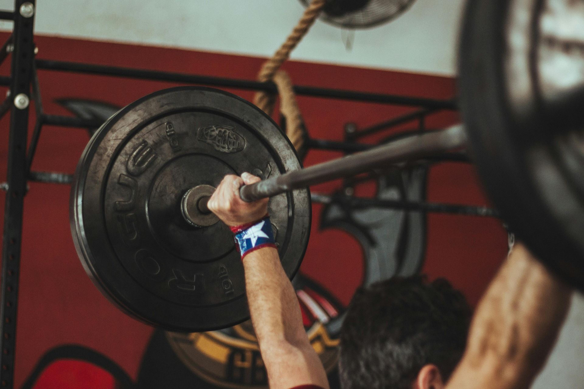 Gewichtheffer tilt een halter boven zijn hoofd in een sportschool; de arm met de blauwe polsband is bedekt.