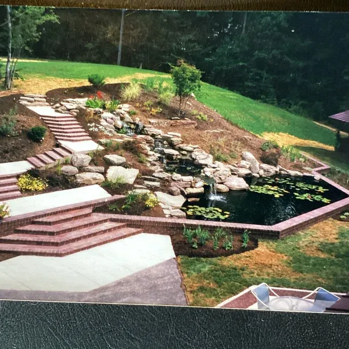 Landscaped yard with a tiered stone staircase leading to a tiered rock waterfall flowing into a dark pond with lilies.