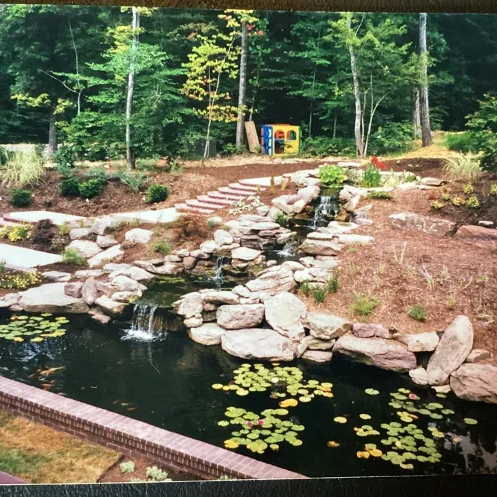 A tiered rock waterfall flows into a pond with lily pads, framed by a brick border in a wooded backyard setting.