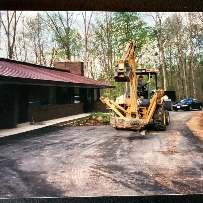 A backhoe lifts a large stone near a house with a brown metal roof and dark siding on a paved driveway.